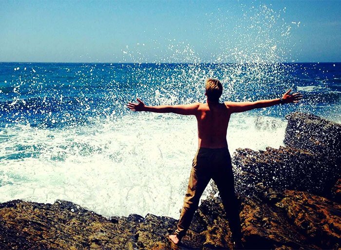 Man standing with arms wide open on edge of rocks in front of crashing waves